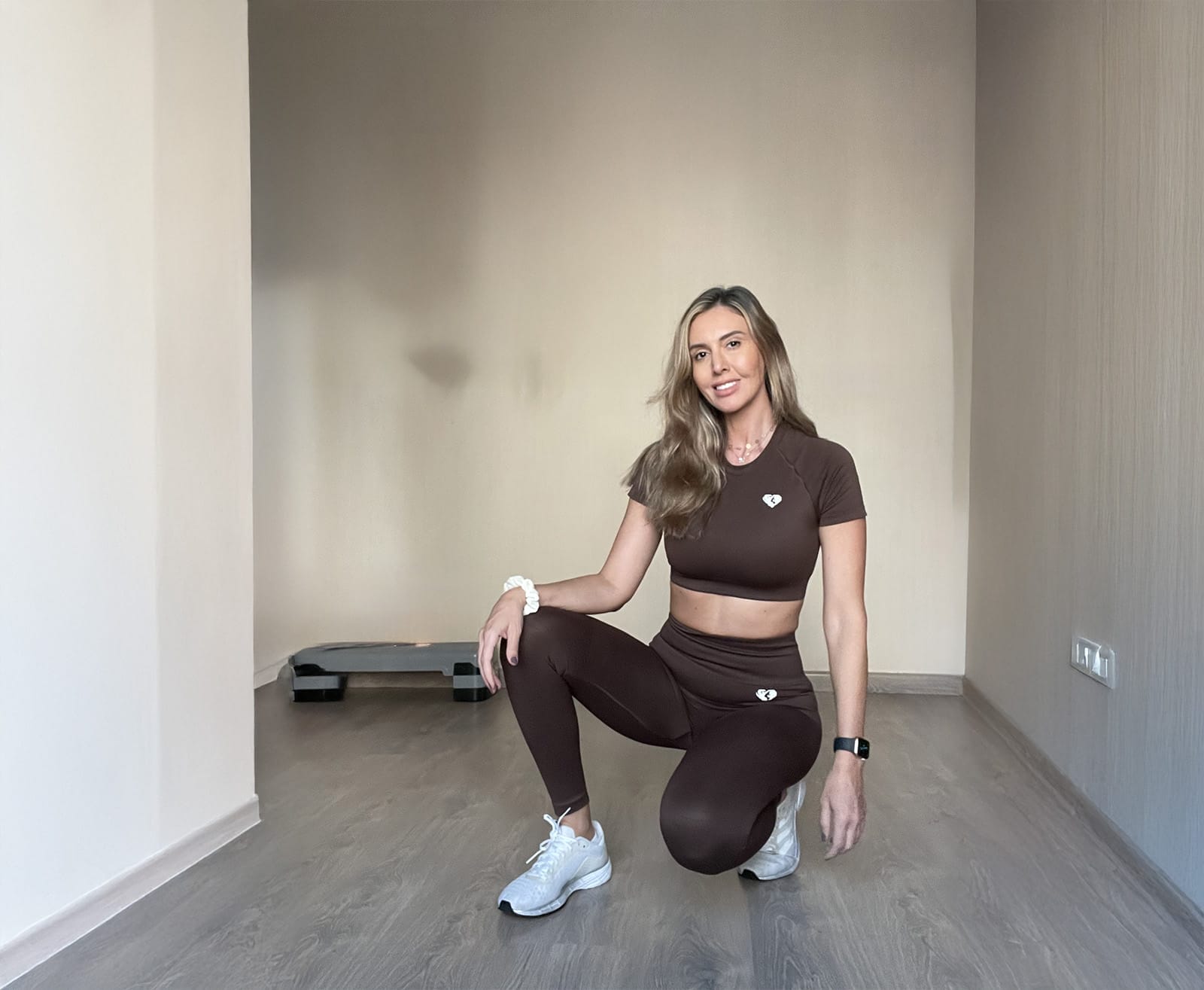 A woman with long blonde hair is kneeling and smiling in a minimal, light-colored room. She wears a matching brown athletic set, white sneakers, a white scrunchie, and a black smartwatch. A small step platform is visible in the back left corner.